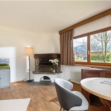 A cozy living space with a fireplace, a table, and modern furniture. Large windows let in plenty of daylight and offer a view of nature.