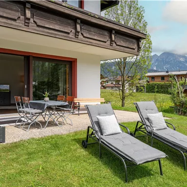 A modern residential building with a cozy outdoor area. Two loungers are placed in the grass, and mountains are visible in the background.