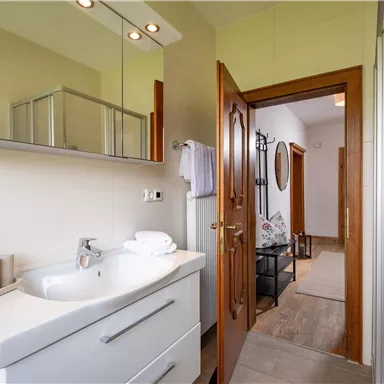 A modern bathroom with a bright vanity and a large mirror. In the background, a wooden slatted door is visible, leading to a hallway.