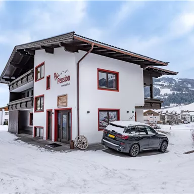 A modern house in the snow with a gray car in front of it. The surroundings are wintry with snow-covered mountains in the background.