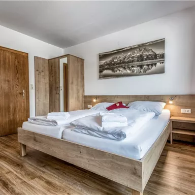A modern bedroom with a large double bed and wooden decor. A landscape painting hangs on the wall, and there is a wardrobe as well as a table.