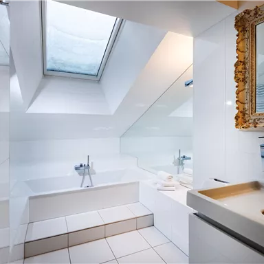 Ein modernes Badezimmer mit einem großen Fenster und einer Badewanne. Die Wände und Fliesen sind in hellen Farben gehalten, was den Raum offen und einladend wirken lässt.