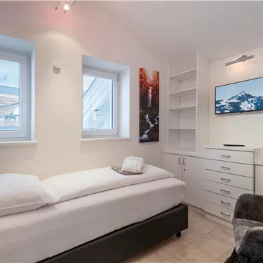 Ein kleines, modernes Schlafzimmer mit einem Einzelbett und einem Fernseher an der Wand. Es gibt Regale und große Fenster, die viel Licht reinlassen.