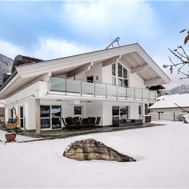 Ein modernes Haus im Schnee mit großen Fenstern und einem Balkon. Im Hintergrund sind schneebedeckte Berge zu sehen.