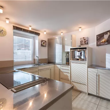 Eine moderne Küche mit hellen Möbeln und grauen Oberflächen. Große Fenster sorgen für viel Tageslicht.