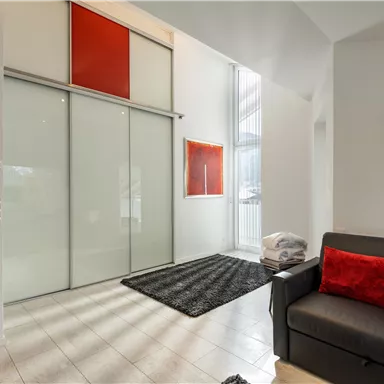 Ein modernes Wohnzimmer mit einem grauen Sofa und roten Kissen. Große Fenster und eine helle, minimalistische Einrichtung prägen den Raum.