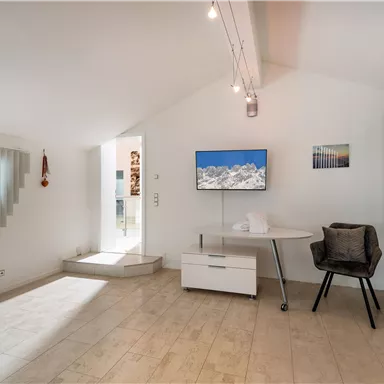 Ein modernes Zimmer mit schrägen Wänden und Holzfußboden. Es gibt einen Schreibtisch, einen Fernseher und große Fenster mit horizontalen Jalousien.