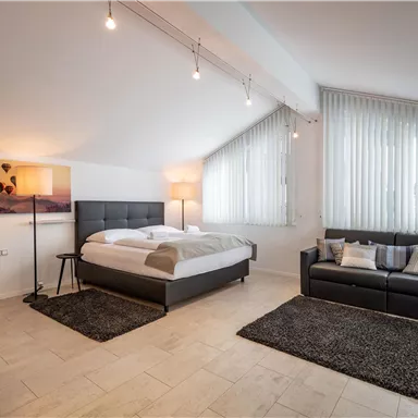 Ein modernes Schlafzimmer mit einem großen Bett, einem Sofa und einem hellen, einladenden Ambiente. Große Fenster sorgen für viel Tageslicht.