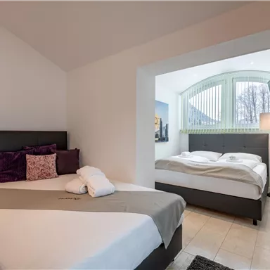 Ein modernes Schlafzimmer mit zwei Betten und neutralen Farben. Große Fenster sorgen für viel Tageslicht.