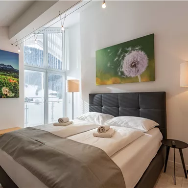 Ein helles Schlafzimmer mit einem Doppelbett und zwei Handtüchern. An den Wänden hängen bunte Bilder, und es gibt eine große Fensterfront.