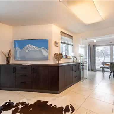 Ein modernes Wohnzimmer mit einem großen Fernseher und einem Blick auf die Berge. Der Boden ist mit Fliesen und einem Teppich versehen, und es gibt einen Esstisch im Hintergrund.