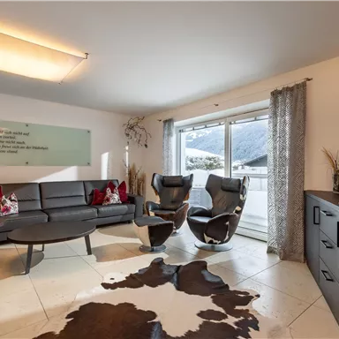 Ein modernes Wohnzimmer mit einem stilvollen Sofa und zwei bequemen Sesseln. Große Fenster bieten einen Blick auf die umliegende Landschaft.