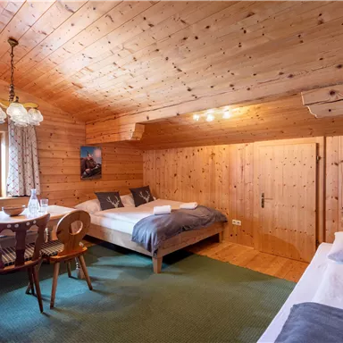 Ein gemütliches Zimmer aus Holz mit zwei Betten und einem Esstisch. Große Fenster lassen viel Licht herein und bieten einen schönen Blick auf die Umgebung.