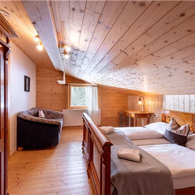 Ein gemütliches Dachzimmer mit Holzverkleidung, zwei Betten und einem kleinen Sitzbereich. Große Fenster sorgen für viel Tageslicht und einen Blick nach draußen.