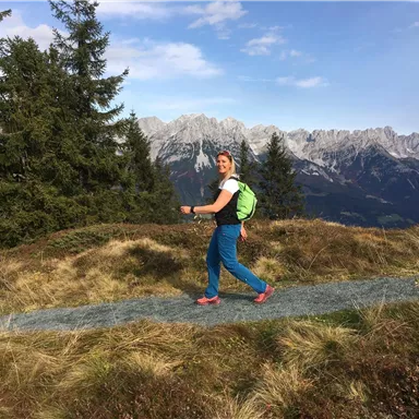A person is hiking on a narrow path in the mountains. In the background, tall mountains and a blue sky can be seen.