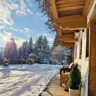 A cozy terrace of a house in the snow with a view of snow-covered trees. The sky is clear and the sun is shining.