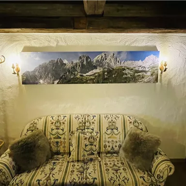 A cozy living room with a beautiful sofa and a landscape painting of the mountains on the wall. The walls are rustic and warmly lit.