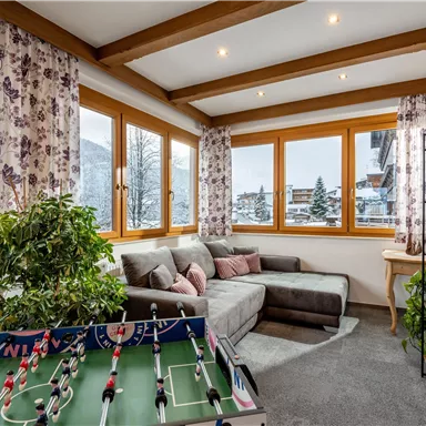 A cozy living space with large windows and a view of snow-covered mountains. In the foreground, there is a foosball table and a comfortable couch with decorative cushions.