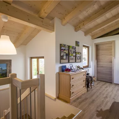 A bright hallway with a wooden ceiling and large windows. Pictures hang on the wall, and a piece of furniture with various items stands there.