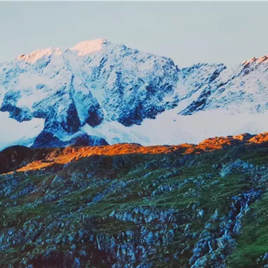 An impressive mountain landscape with snow-capped peaks and green slopes. The dawn illuminates the landscape in warm colors.
