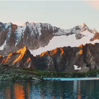 An impressive mountain landscape with snow-capped peaks and warm light. In the foreground, a calm lake reflects the mountains.
