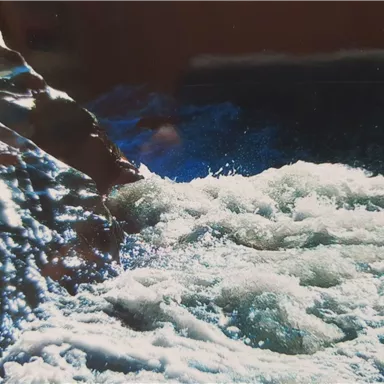 A rushing flow of water meets a rocky shoreline. The water shimmers in various shades of blue and green.