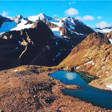 An impressive mountain landscape with snow-covered peaks and the clear blue of a small lake. The sky is bright blue with a few clouds.