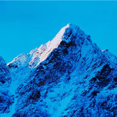 A snow-covered mountain peak under a clear blue sky. The summit is illuminated by sunlight.