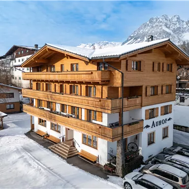 A charming wooden house in the snow with multiple floors and balconies. In the background, snow-covered mountains and a clear sky are visible.