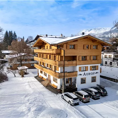 A cozy wooden building in the snow, surrounded by trees. There are several cars in the foreground and a mountain landscape in the background.