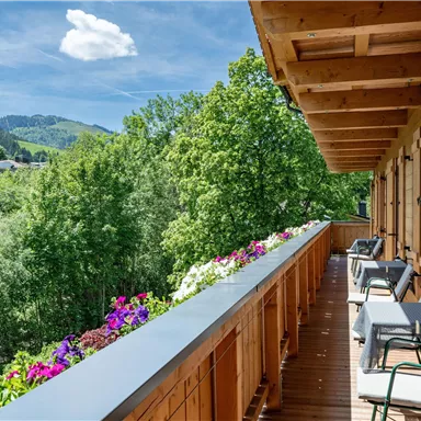 Ein schöner Balkon mit Holzverkleidung und Aussicht auf grüne Bäume und Berge. Blumen schmücken das Geländer und laden zum Entspannen ein.