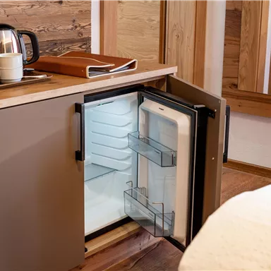 Ein kleiner Kühlschrank in einem gemütlichen Raum. Neben dem Kühlschrank steht eine Teekanne und eine Schreibunterlage.