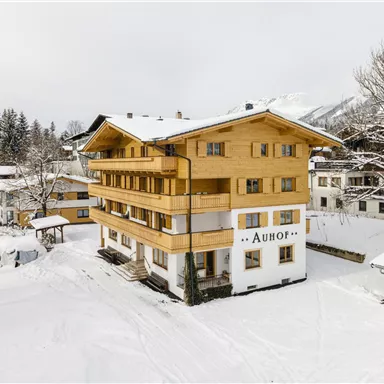 Ein Holzhaus im Schnee mit mehreren Stockwerken. Umgeben von verschneiten Bäumen und anderen Gebäuden.