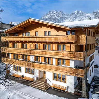 A cozy wooden house in the snow with several balconies. In the background, majestic mountains and a clear blue sky are visible.