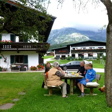 Eine Gruppe von fünf Personen sitzt an einem Tisch im Freien, umgeben von einem schönen Garten. Im Hintergrund sind Berge und traditionelle bayerische Häuser zu sehen.