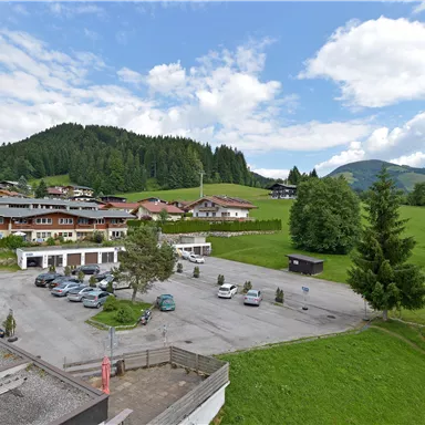 A peaceful landscape with green meadows and mountains in the background. Several houses and a parking lot are visible.