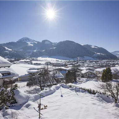 A winter landscape with snow-covered roofs and mountains in the background. The sun shines in the clear blue sky.