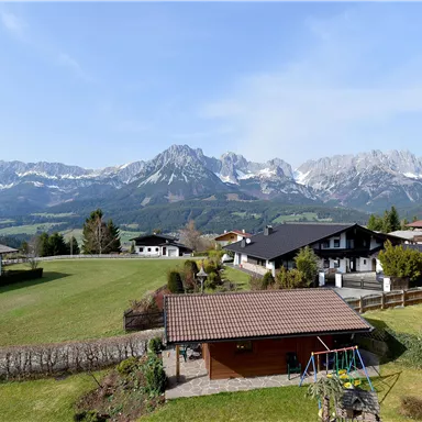 Eine malerische Landschaft mit schneebedeckten Bergen im Hintergrund. Im Vordergrund sind Wiesen und traditionelle Häuser zu sehen.