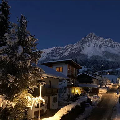 Eine winterliche Landschaft bei Nacht mit schneebedeckten Bergen und beleuchteten Häusern. Der Himmel ist dunkelblau und die Umgebung wirkt ruhig und friedlich.