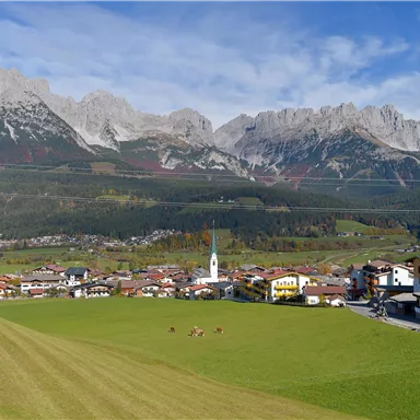 A picturesque mountain landscape with majestic peaks and a small village in the foreground. The meadows are green and there is a clear blue sky.