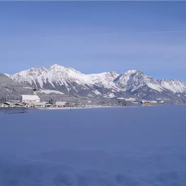 Eine verschneite Landschaft mit Bergen im Hintergrund. Ein ruhiges Dorf ist am Fuße der Berge sichtbar.