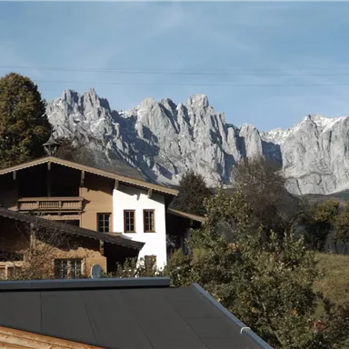 A picturesque mountain landscape with snow-capped peaks in the background. In the foreground, traditional houses and green trees can be seen.