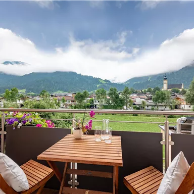 A cozy balcony with a small table and chairs. In the background, green mountains and a charming village can be seen under a cloudy sky.