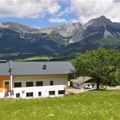 A modern house sits on a green meadow with a view of majestic mountains. The sky is clear and the surroundings are rural and idyllic.