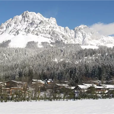 Eine schneebedeckte Berglandschaft mit hohen Gipfeln und einem klaren blauen Himmel. Im Vordergrund sind mehrere Häuser und Bäume im Schnee zu sehen.