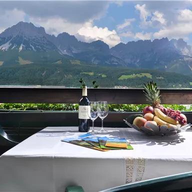 A beautiful table with fruit and wine is on a terrace overlooking majestic mountains. The clouds in the sky give the picture a relaxed atmosphere.