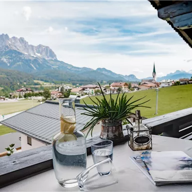 A view from a balcony overlooking a picturesque mountain landscape. On the table are a water pitcher and glasses, surrounded by a green meadow.