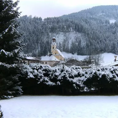Eine verschneite Landschaft mit Tannenbäumen und einem kleinen Dorf. Im Hintergrund ist eine Kirche sichtbar, umgeben von schneebedeckten Bergen.