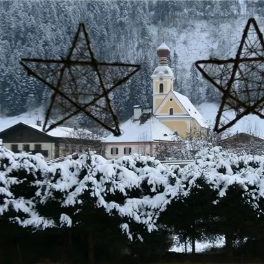 Eine verschneite Landschaft mit einem charmanten Dorf und einer Kirche im Hintergrund. Der Blick ist durch zwei dekorative Sterne gerahmt.