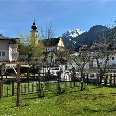 Ein idyllisches Dorf mit schönen Häusern und einem Kirchturm. Im Hintergrund sind schneebedeckte Berge und ein klarer blauer Himmel zu sehen.